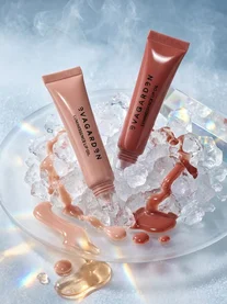 Twee tubes EVAGARDEN Luminessence Lip Oil in zachtroze en nude bruin liggend op een bed van glanzende ijsblokjes.